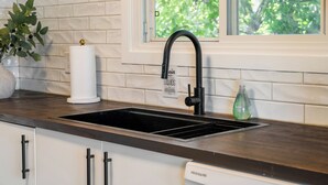 Una cocina con grifo negro, azulejos blancos, una ventana con vistas a árboles, una planta en maceta y una botella de jabón para manos.