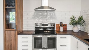 Una cocina con estufa de acero inoxidable, armarios blancos y un mueble de madera con puertas de cristal.