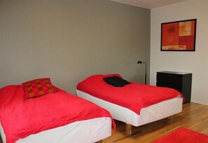 Une chambre avec deux lits simples, chacun recouvert d’une couverture rouge et agrémenté d’oreillers blancs. On y trouve une table de chevet noire et un tapis rouge sur le sol.