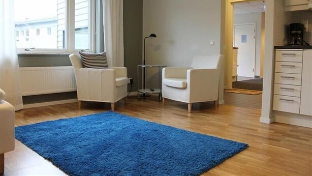 A compact living space with a blue rug, two white armchairs, a small table, and a kitchenette area. - Hotel Torslanda Studios (Torslanda)