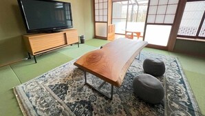 A traditional Japanese-style room with tatami flooring, a low wooden table, two cushions, a television on a wooden stand, and sliding doors with a view of the outside. - Assi (Yufu)