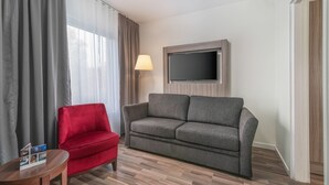 A hotel room with a grey sofa, a red armchair, a flat-screen TV mounted on the wall, and a lamp beside the sofa. - First Hotel Oxelösund (Oxelosund)