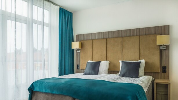 A hotel room with a large bed, two bedside lamps, a headboard, and a view of the city through large windows. - First Hotel Oxelösund (Oxelosund)