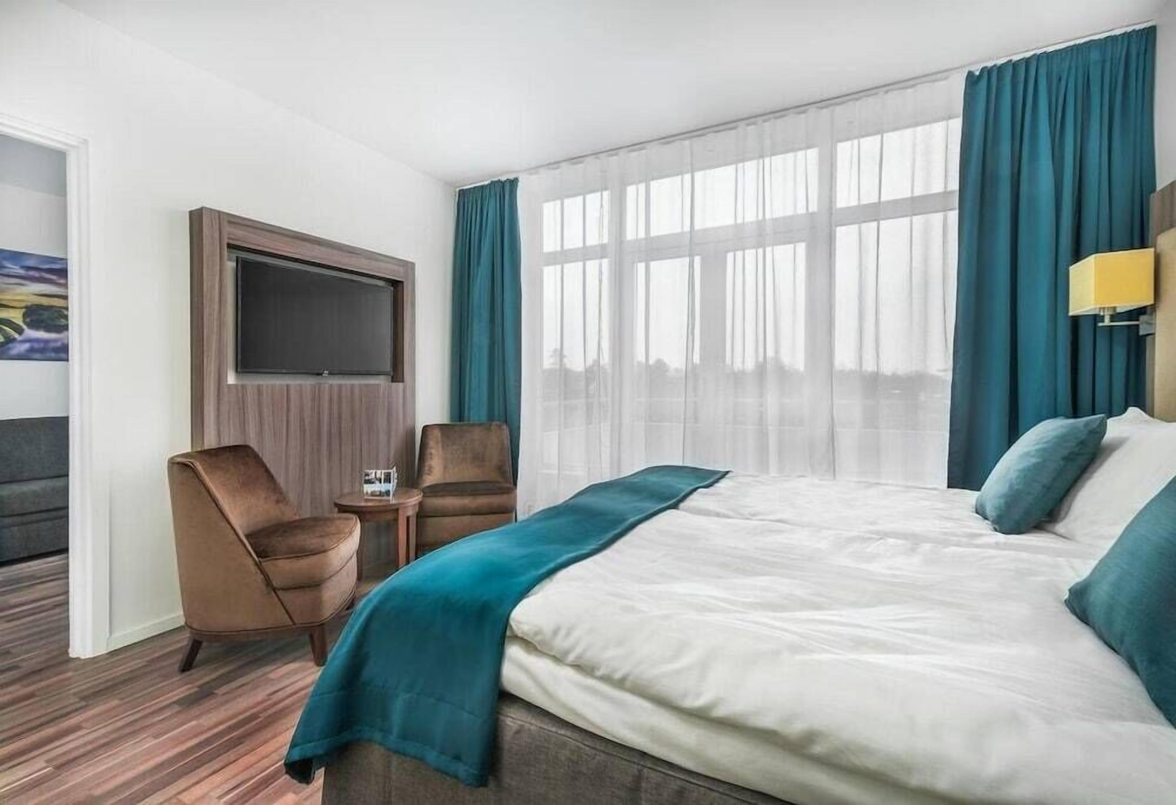 A hotel room with a large bed, two chairs, a TV, and a window with curtains.