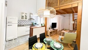 Cocina compacta con armarios blancos, microondas y lavabo. Hay una zona de comedor con una mesa para dos, sillas y una lámpara colgante.