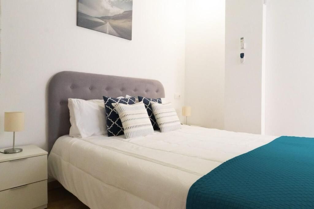 Una cama bien hecha con cabecera gris y ropa de cama blanca, una mesita de noche con lámpara y un cuadro en la pared.