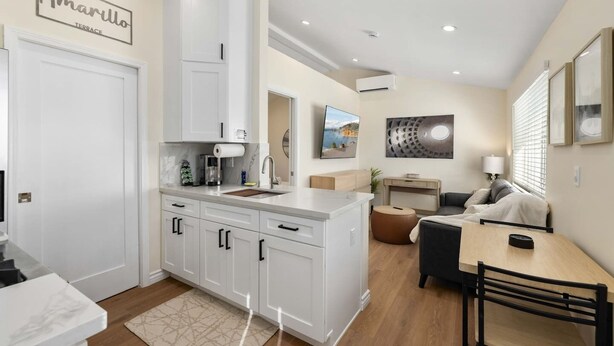 A modern kitchen with white cabinets, a built-in oven, and a large island with a sink. There is a dining area with a wooden table and chairs, and a living space with a sofa and a TV. - Luxury 1BR Suite Kitchen, Fireplace, Laundry, BBQ (Simi Valley)