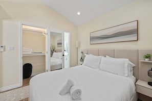 Habitación de hotel con una cama grande, una mesita de noche, un armario con ropa colgada y una puerta que da a otra habitación.