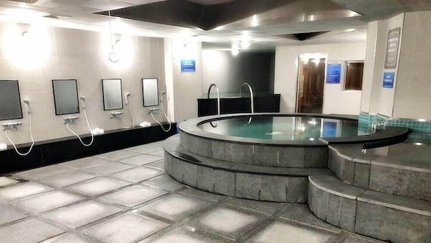 Un espace piscine intérieur moderne, doté d’une paroi vitrée, d’un revêtement de sol en pierre et de plusieurs douches.