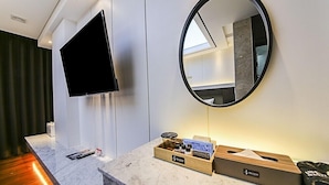 Une chambre d’hôtel dotée d’un comptoir en marbre, d’un téléviseur à écran plat fixé au mur, d’un miroir rond et d’une table d’appoint en bois avec des produits de première nécessité.