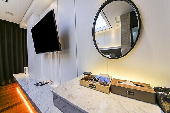 Ett hotellrum med en marmorskiva, en platt-TV som är monterad på väggen, en rund spegel och ett litet sidobord i trä med diverse bekvämligheter.
