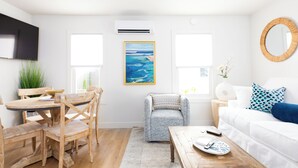 A modern living room with a dining area, featuring a wooden dining table and chairs, a grey armchair, a white sofa with blue cushions, and a large painting on the wall.