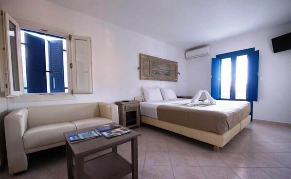 A small, clean room with a bed, a sofa, a nightstand, and a window with blue shutters. - Amorgion Hotel (Amorgos)