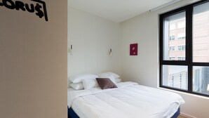 A bedroom with a bed, white walls, a window with a view of buildings, and a small red decoration on the wall. - Yust Antwerpen (Antwerp)