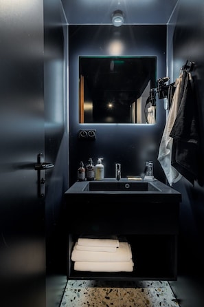 Une salle de bain moderne aux couleurs sombres, dotée d’un grand miroir, d’un lavabo avec des articles de toilette et d’un porte-serviettes.