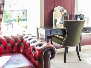 A room with a red leather sofa, a green armchair, a desk with a mirror, and a window with a view of greenery. - The Dirty Bottles Inn (Alnwick)