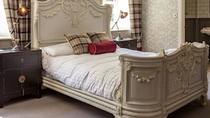Una cama clásica con cabecera y piecera decorados, una mesita de noche con lámpara y otra mesita con lámpara.