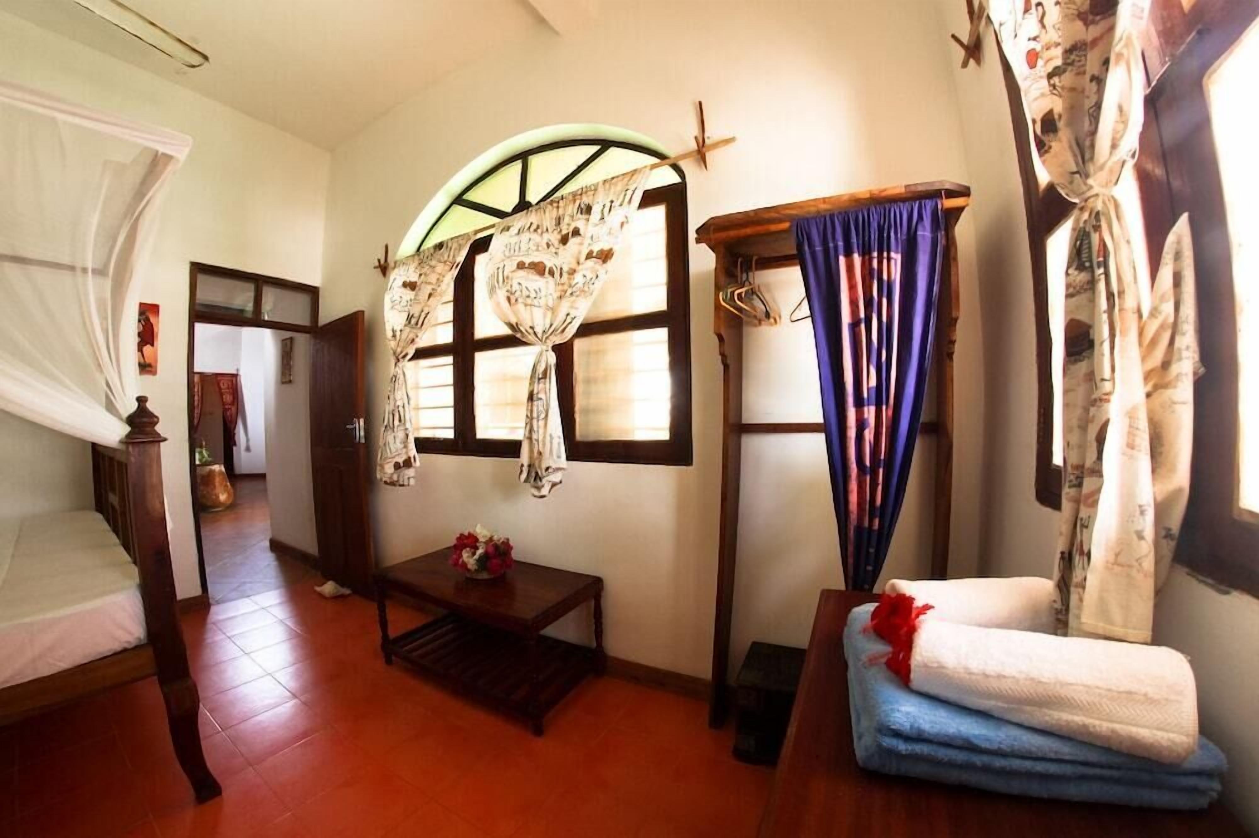 A room with a bed, a small table with a vase, a window with curtains, and a door.