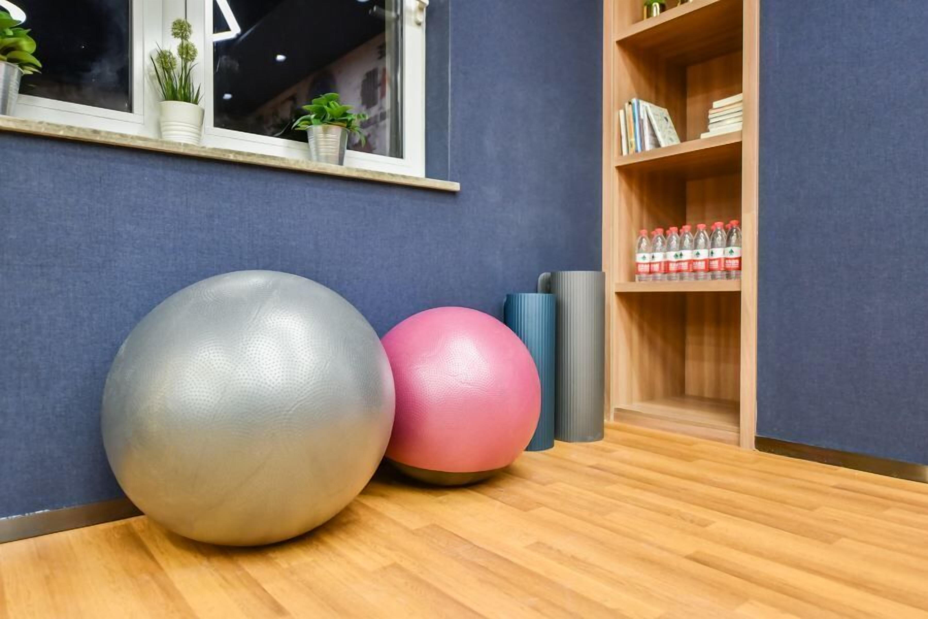 A room with exercise balls, a window with plants, and a shelf with books and water bottles.