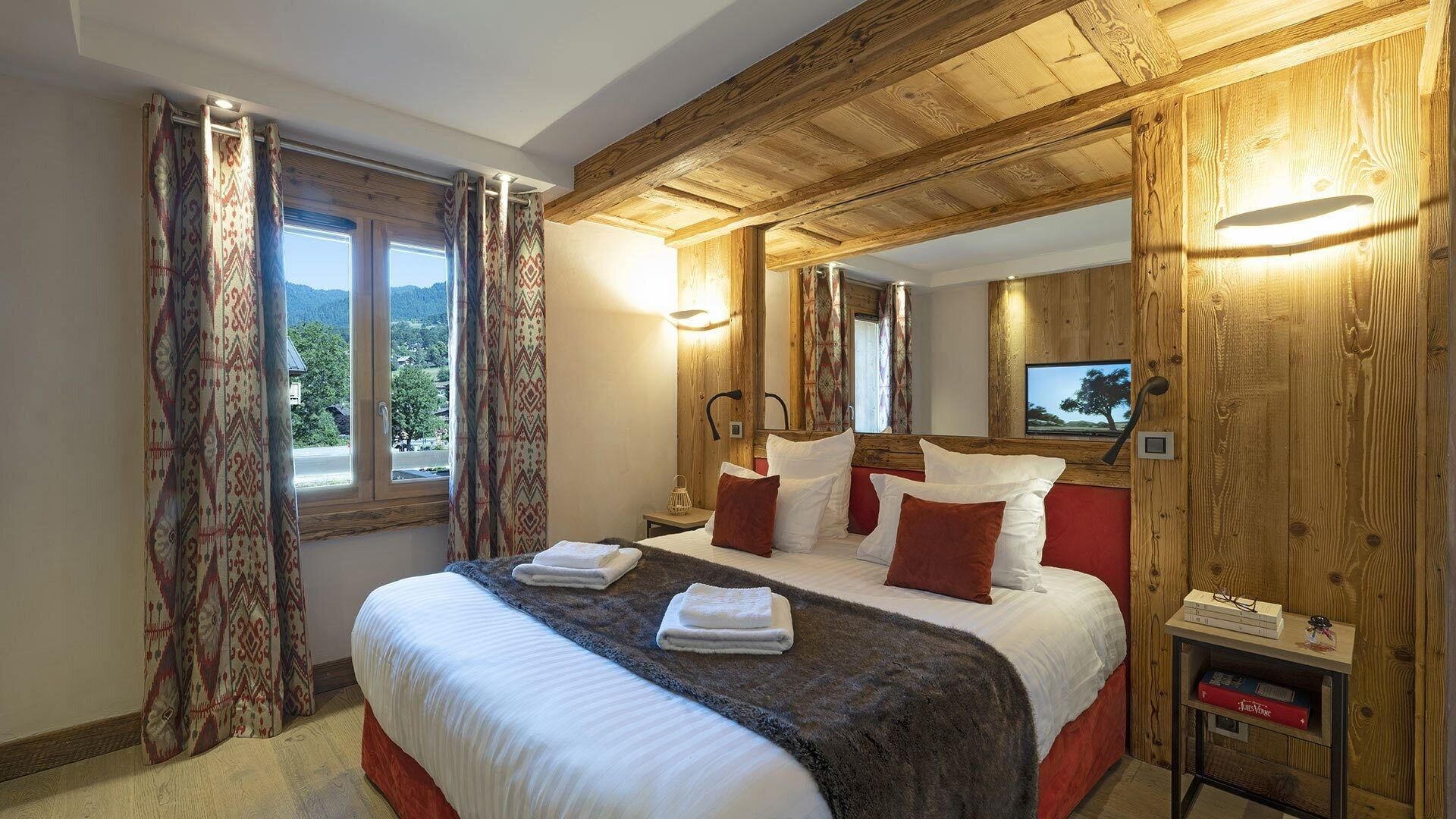 Une chambre avec un plafond en bois, un lit avec une literie blanche et rouge, une table de chevet avec une lampe, et une fenêtre avec des rideaux.