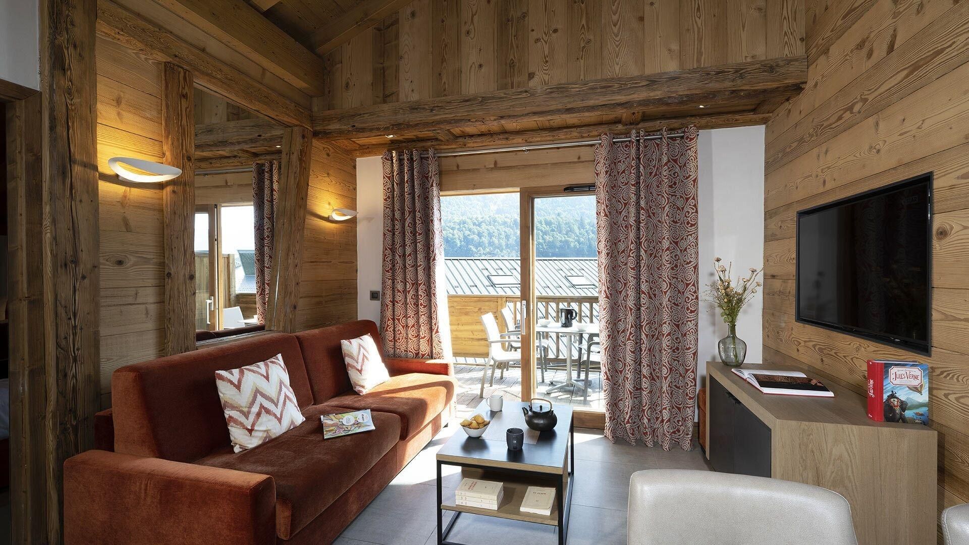 A cozy room with a wooden interior, a sofa, a coffee table, a TV, and a balcony with patterned curtains.