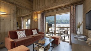 A cozy living room with a wooden interior, a sofa, a coffee table, and a view of the outdoors. - MGM Hôtels et Résidences - Residence Alexane (Samoëns)