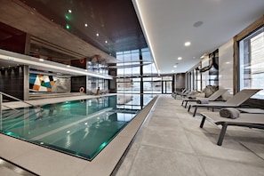 A modern indoor swimming pool with lounge chairs and a large window providing a view of the outside. - MGM Hôtels et Résidences - Residence Alexane (Samoëns)