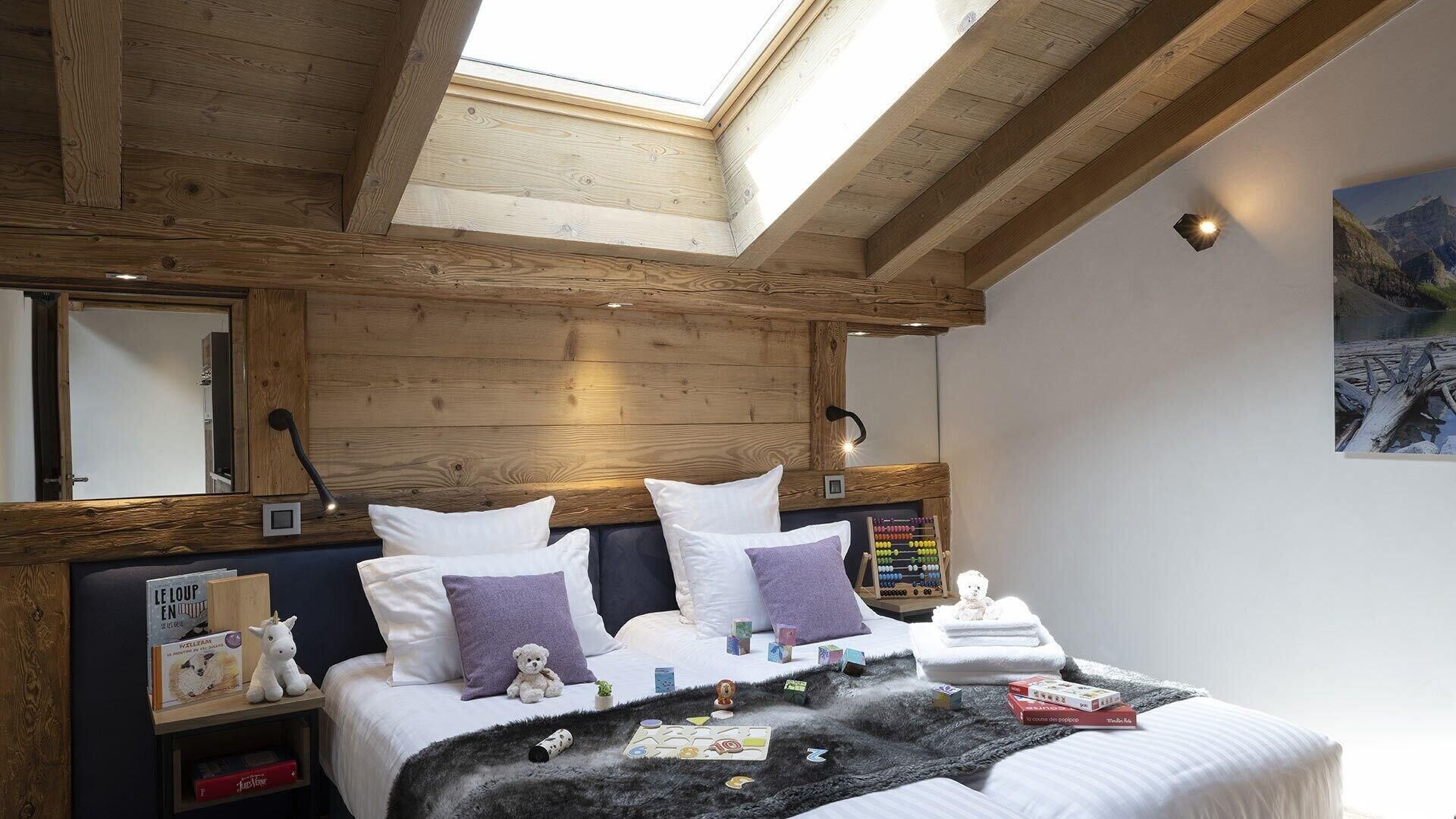A bedroom with a sloped ceiling, wooden beams, a skylight, a bed with pillows, a nightstand, a teddy bear, and a small table with books and a lamp.