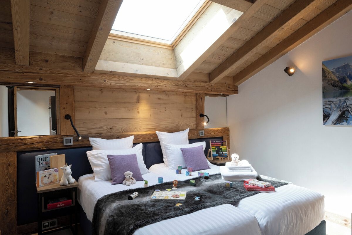 A bedroom with a skylight, wooden beams, a bed with white bedding, a nightstand with a lamp, and a wall-mounted clock.