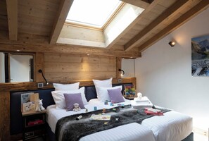 A bedroom with a skylight, wooden beams, a bed with white bedding, a nightstand with a lamp, and a wall-mounted clock. - MGM Hôtels et Résidences - Residence Alexane (Samoëns)