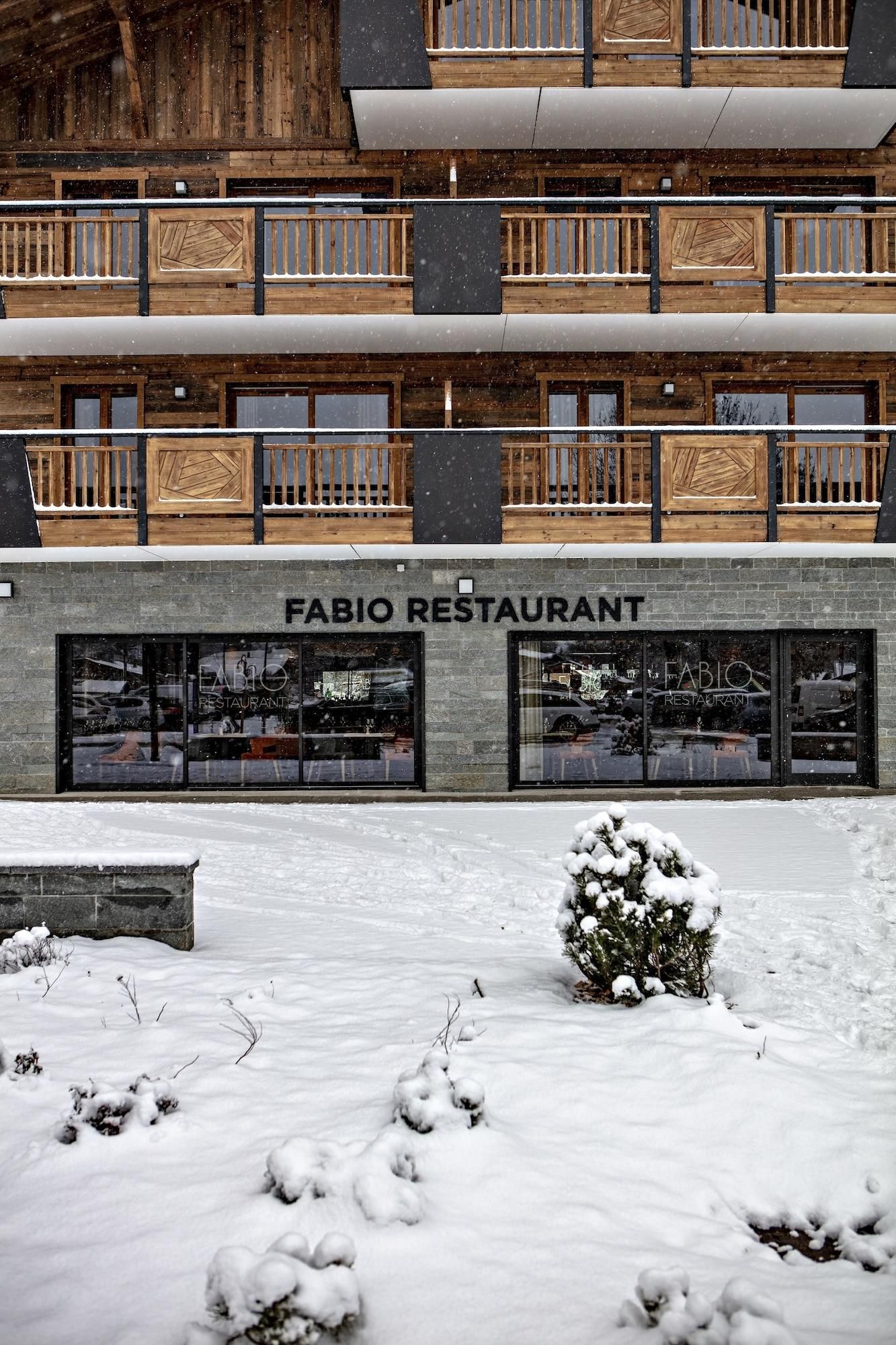 Ein zweistöckiges Gebäude mit einer Holzfassade und Balkonen. Im Erdgeschoss befinden sich große Fenster und eine Schilder mit der Aufschrift 'FABIO RESTAURANT'. Schnee bedeckt den Boden und ein kleiner Strauch ist zu sehen.
