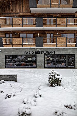 A two-story building with a wooden facade and balconies. The ground floor has large windows and a sign that reads 'FABIO RESTAURANT.' Snow covers the ground and a small bush. - MGM Hôtels et Résidences - Residence Alexane (Samoëns)