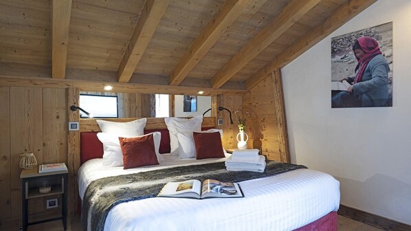 A cozy bedroom with a wooden ceiling, a bed with white and red pillows, a bedside table with a lamp, and a framed picture on the wall. - MGM Hôtels et Résidences - Residence Alexane (Samoëns)