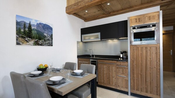 A modern kitchen with wooden cabinets, a built-in oven, and a dining area with a table set for two. - MGM Hôtels et Résidences - Residence Alexane (Samoëns)