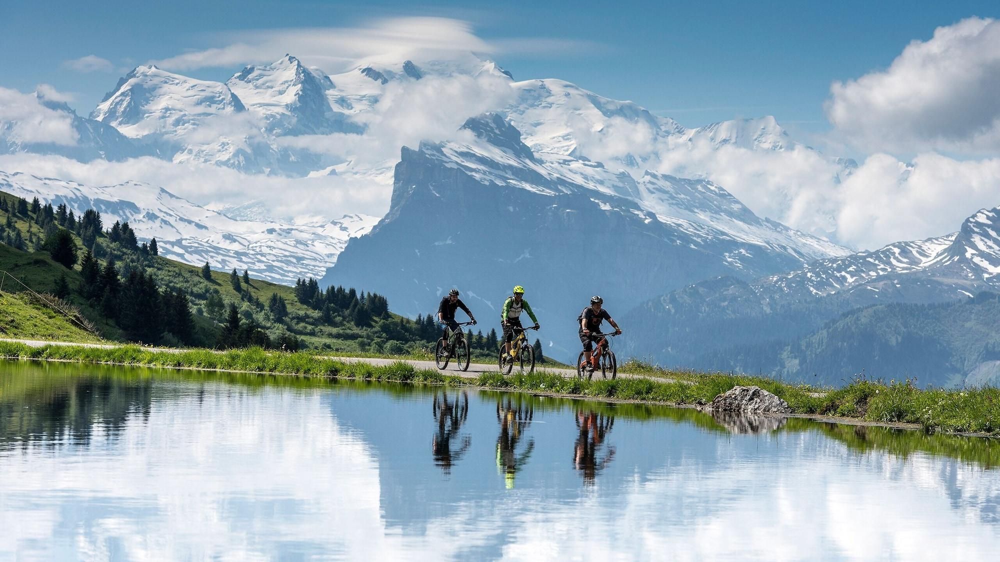 Un paysage de montagne avec des cyclistes sur un sentier, près d’une étendue d’eau qui reflète la lumière.