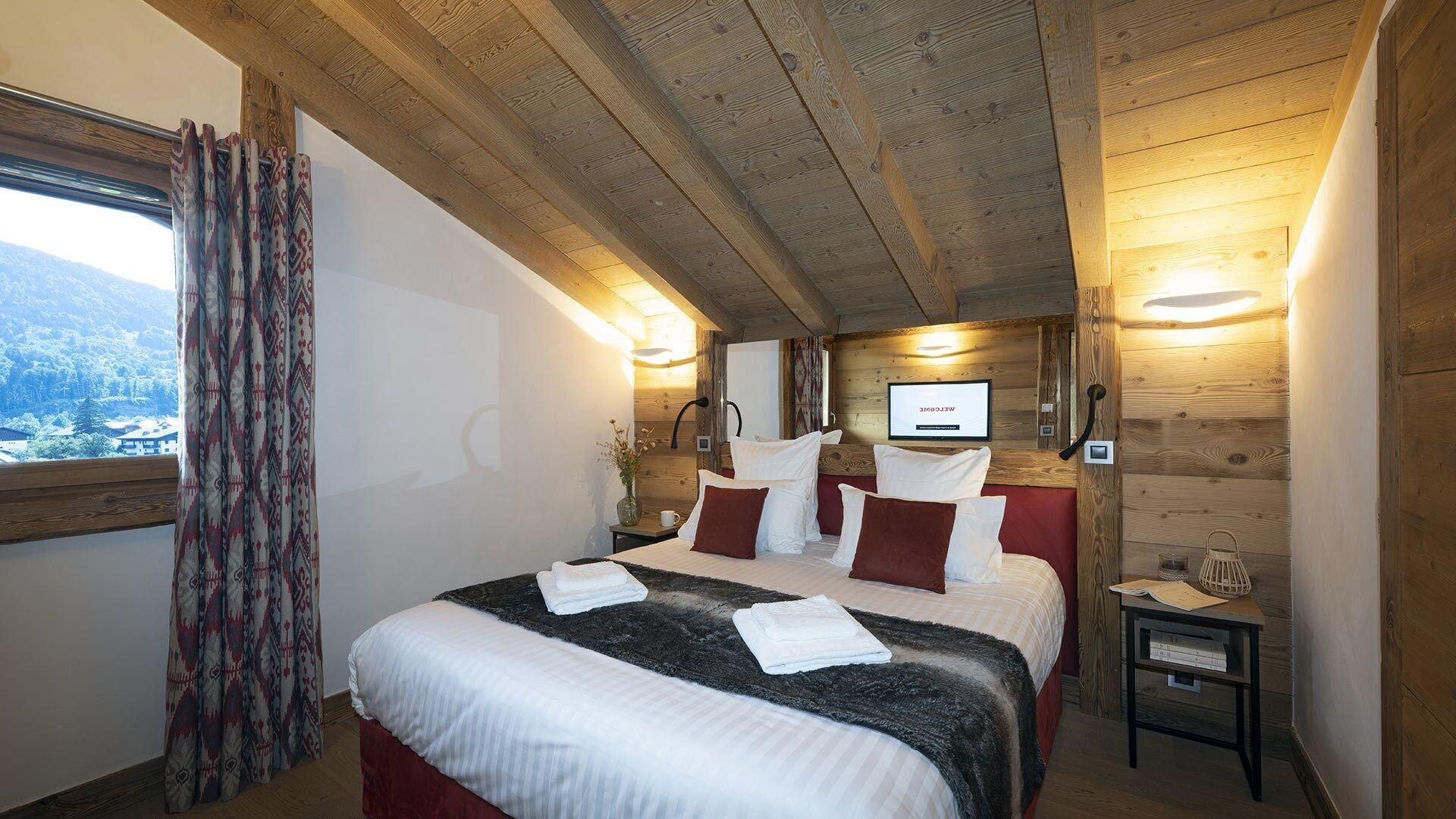 A cozy room with a bed, a small table, a nightstand, a window with curtains, and a wooden ceiling.