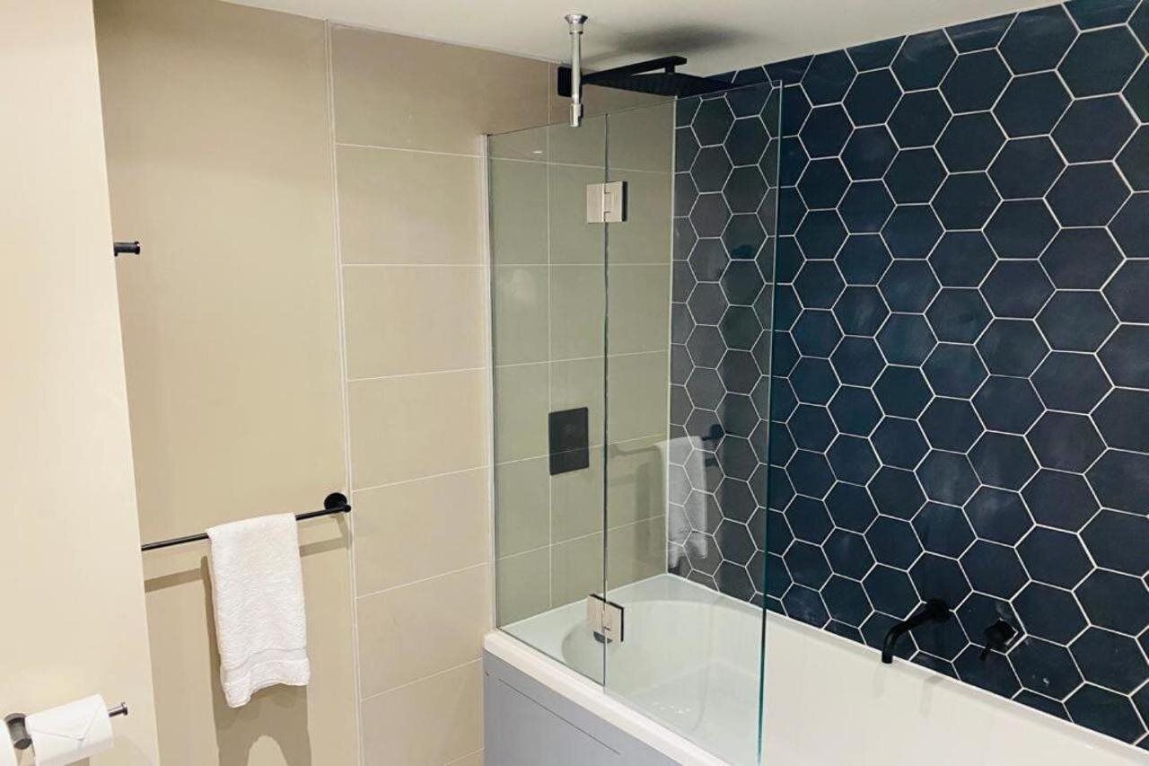 A modern bathroom with a glass-enclosed shower, a white bathtub, and a hexagonal tile pattern on the wall.