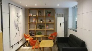 A modern waiting area with a black sofa, wooden chairs, a small table, and a bookshelf with plants and books. - 7 Days Inn Premium Beijing Dongzhimen Airport Express Station (Beijing)