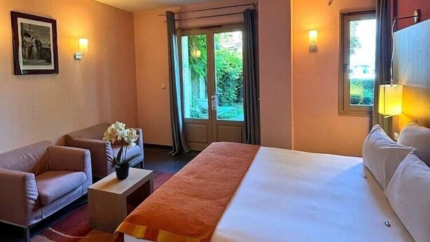 Una habitación de hotel con una cama grande, un sofá, una mesita con un jarrón y una ventana con cortinas.