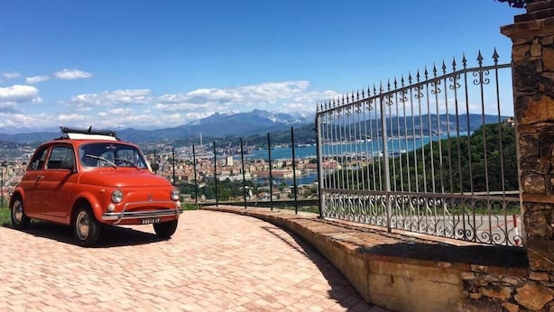 Un coche clásico rojo aparcado en una zona pavimentada, con vistas a un pueblo costero y montañas al fondo.
