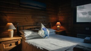 Une chambre chaleureuse aménagée dans un chalet en rondins, avec un lit, deux lampes de chevet, une tête de lit en bois et une fenêtre donnant sur le ciel.