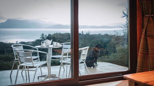 Une pièce dotée d’une grande fenêtre donnant sur un paysage magnifique, avec une étendue d’eau et des montagnes en toile de fond. À l’intérieur, on trouve une table avec des chaises et un chien assis près de la fenêtre.