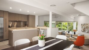 A modern kitchen with a central island, white chairs, a dining table with a vase of flowers, and a living area with a sofa and a coffee table. - Parkview on Sunshine 2 Sunshine Beach (Sunshine Beach)