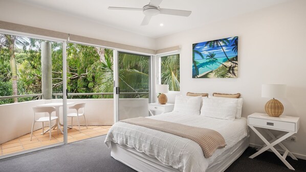 A bedroom with a large bed, a ceiling fan, a nightstand with a lamp, a painting of a beach scene, and a balcony with a chair and table.