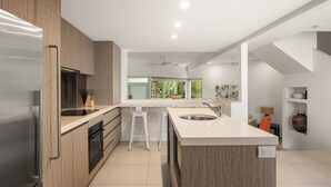 A modern kitchen with a central island, wooden cabinets, and a large window providing a view of greenery. - Parkview on Sunshine 2 Sunshine Beach (Sunshine Beach)