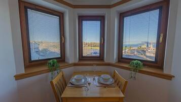 A dining area with a table set for two, flanked by three windows with blinds, each offering a view of a construction site.