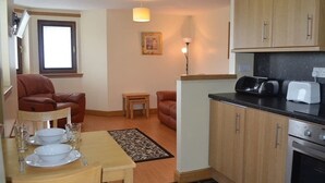 A compact kitchen with wooden cabinets, a built-in oven, and a dining area with a table set for two. - Quay Head View (Stranraer)
