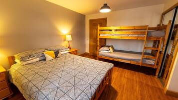 A bedroom with a large bed, a smaller bed, and a bunk bed. There is a nightstand with a lamp, a wooden floor, and a door in the background.