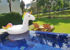 A swimming pool with a unicorn float, two wooden lounge chairs, and a grassy area with trees in the background. - S - Ocean Luxury Villas Danang (Da Nang)