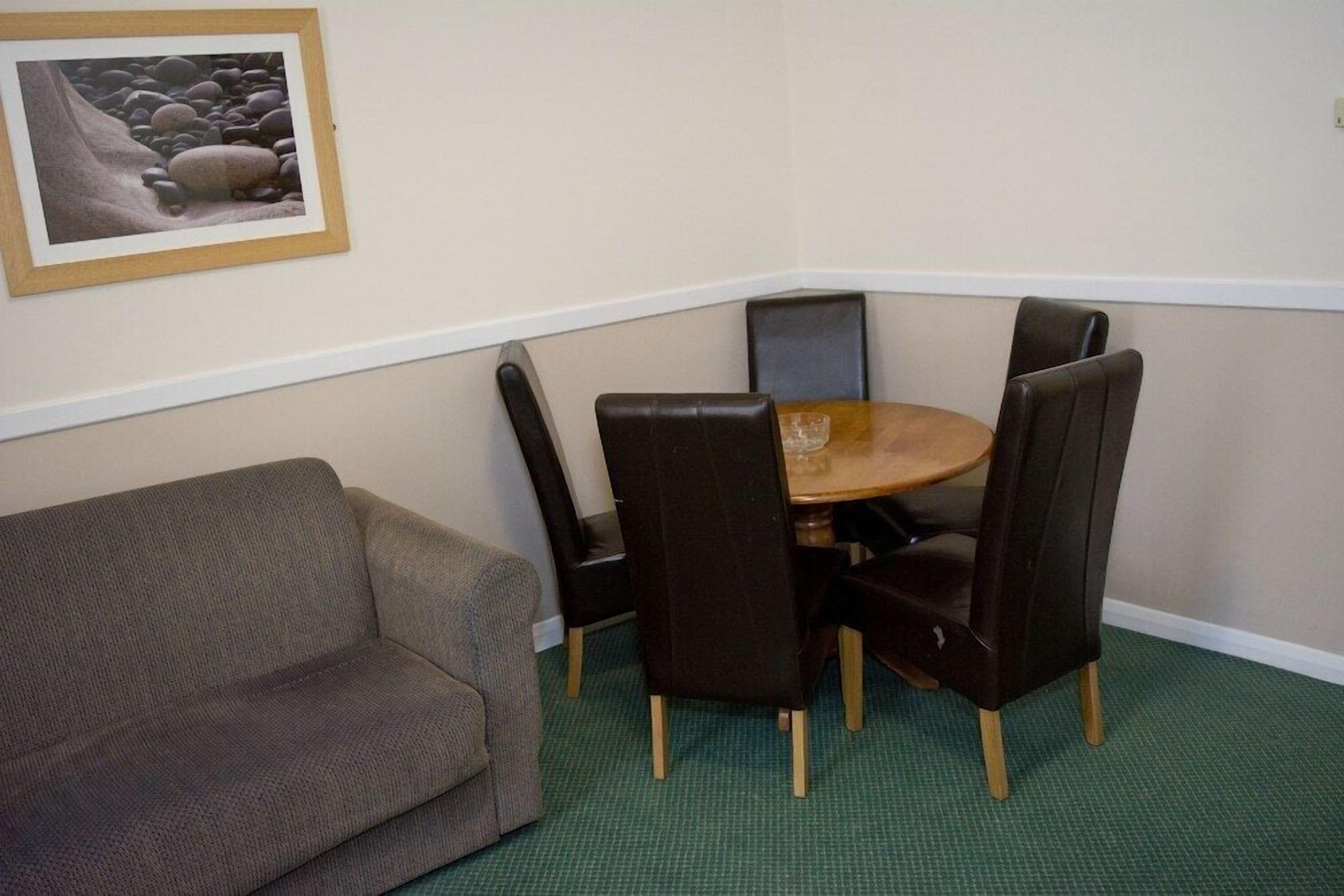 A corner room with a green carpet, a brown sofa, a wooden table, and four black chairs.